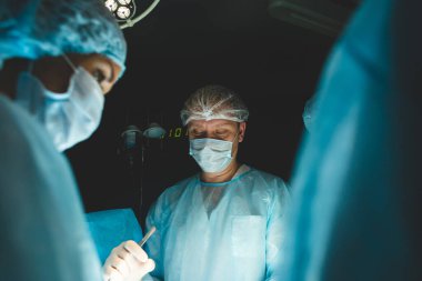 Uluslararası profesyonel bir cerrah, asistan ve anestezi uzmanı genel anestezi altındaki bir hasta üzerinde karmaşık bir operasyon gerçekleştirir. Karanlık atmosfer fotoğrafçılığı teması.
