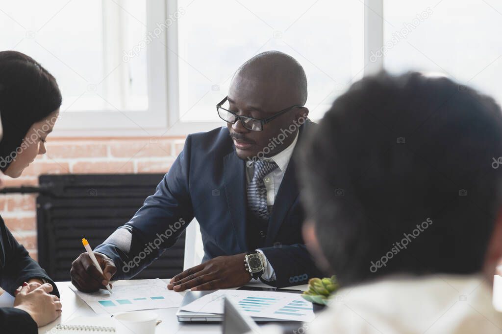 Mature afroamerican businessman to discuss information with a younger colleague. People working and communicating while sitting at the office desk together with colleagues sitting.