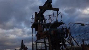 Enayi çubuğu pompalarının silueti çalışıyor ve akşam gökyüzünün zeminine yağ pompalıyor. Petrol kuyusu servisi