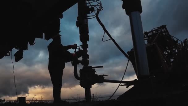 Tulumlu ve kasklı bir çalışan bir petrol kuyusunun onarımı ve bakımını yapıyor. Akşam gökyüzünün arka planında siluet — Stok video