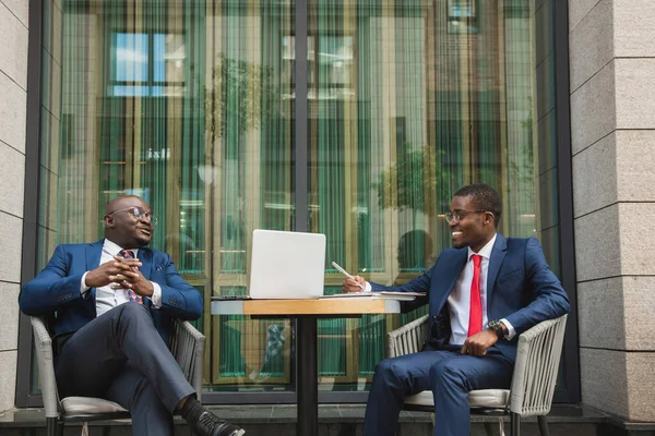 Dos hombres de negocios afroamericanos de piel oscura en trajes y vasos con maletines se sientan en una mesa en un café de la ciudad al aire libre. — Foto de Stock
