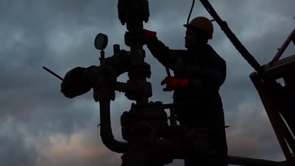 Een medewerker in overalls en een helm is bezig met reparaties en onderhoud van een oliebron. Silhouet op de achtergrond van de avondhemel — Stockvideo