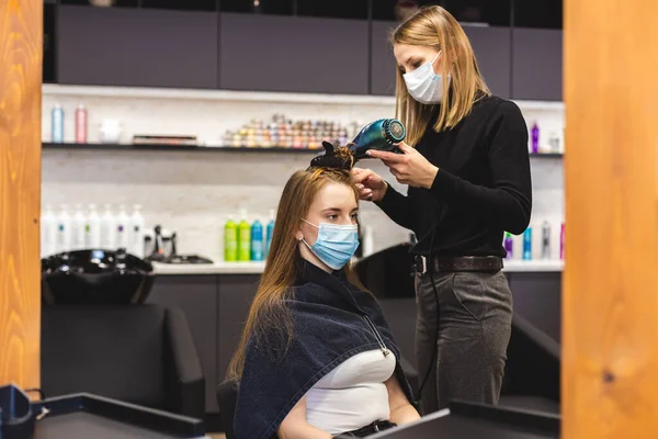 Mistryně žena kadeřnice v lékařské masce suší dívky vlasy s fénem a hřebeny po umytí v salonu krásy. Covid-19 pandemie a opravné prostředky — Stock fotografie