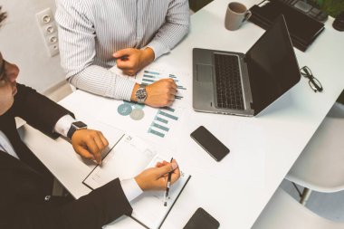 Masada oturan genç bir ofis çalışanı, bilgisayar kullanıyor. İki iş adamı konuşuyor.