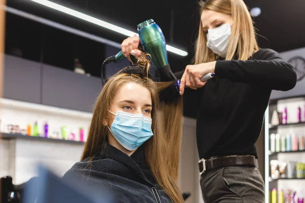 Maître femme coiffeur dans un masque médical sèche les cheveux des filles avec un sèche-cheveux et peignes après le lavage dans un salon de beauté. Covid-19 pandémie et remèdes — Photo