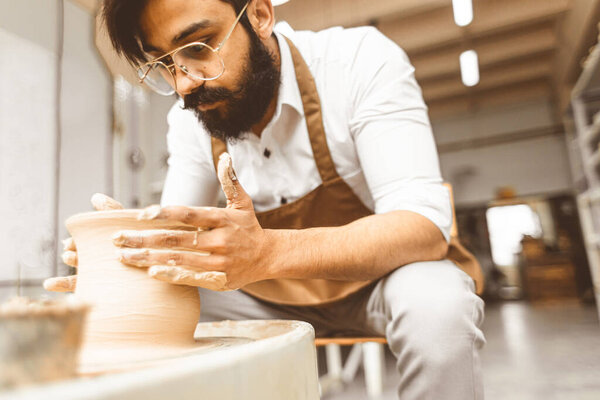 A young male potter is engaged in craft in his workshop on a potters wheel and makes a clay product