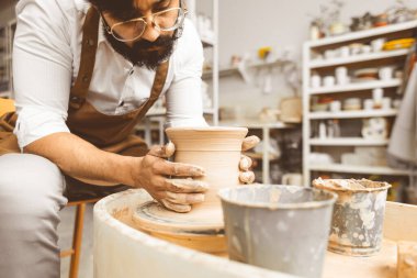 Genç bir erkek çömlekçi atölyesinde çömlekçi çarkında el işi yapıyor ve kilden bir ürün yapıyor.