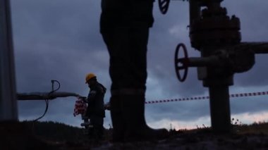 Tulumlu ve kasklı bir çalışan bir petrol kuyusunun onarımı ve bakımını yapıyor. Akşam gökyüzünün arka planında siluet