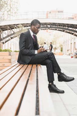 Takım elbiseli, gözlüklü ve kulaklıklı siyahi bir Amerikalı işadamının portresi şehir meydanındaki bir bankta duruyor.