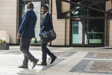 Koyu tenli iki Afro-Amerikalı takım elbiseli ve gözlüklü iş adamı şehirde yürüyüp iş konuşuyorlar.