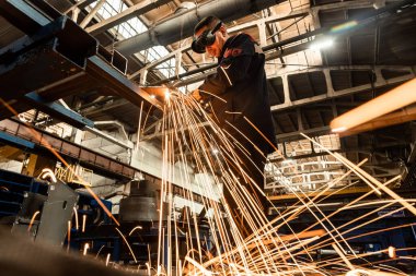 Bir fabrika işçisi, bir üretim hangarının zeminine karşı koruyucu bir maske takan kaynak makinesiyle metal kaynak yapıyor..