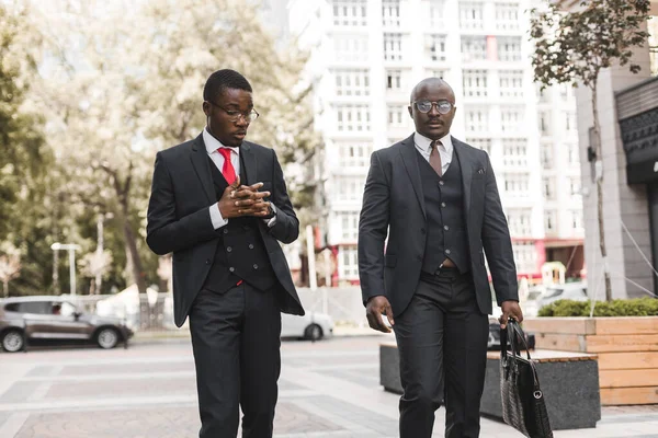 Deux hommes d'affaires afro-américains à la peau foncée en costume et lunettes marchent à l'extérieur dans la ville et discutent affaires — Photo