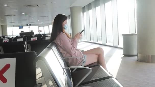 Una giovane donna che indossa una maschera all'aeroporto durante la pandemia di Covid-19 siede su una sedia in una sala di partenza vuota. distanza sociale — Video Stock