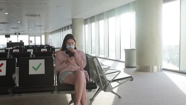 Молодая женщина в маске в аэропорту во время пандемии Ковид-19 сидит в кресле в пустом зале вылета. социальное расстояние — стоковое видео