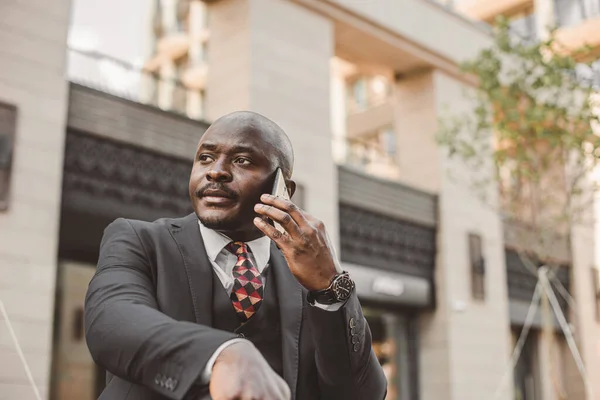 Retrato de empresário negro afro-americano em um terno com óculos e fones de ouvido fica contra o fundo de edifícios da cidade ao ar livre — Fotografia de Stock