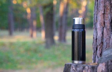 Bir ağaç kütüğünün üzerinde siyah bir termos şişesinin yakın çekimi. Bulanık orman arkaplanı, kopyalama alanı.