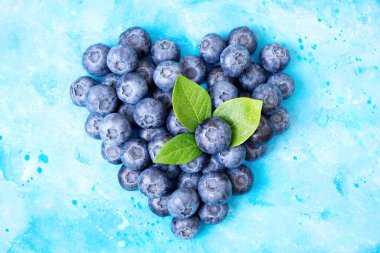 Açık mavi suluboya arka planda taze yaban mersinleri ve yeşil yapraklardan yapılmış kalp sembolü. Sağlıklı beslenme ve bağışıklık güçlendirici kavramı.