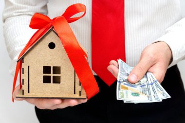 Satıcı bir elinde minyatür ahşap bir ev modeli, diğer elinde de birkaç dolarlık banknot tutuyor. Gayrimenkul alım ve satım kavramı.