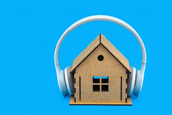 Wireless overhead headphones on a wooden house model isolated on a blue background. Creative apartment noise insulation concept.