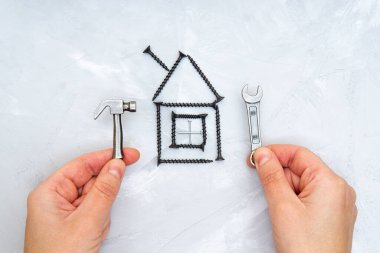 Minyatür çelik bir çekiç tutan eller ve beton bir arka planda siyah renkli vidalardan yapılmış bir ev şeklindeki ingiliz anahtarı. Yaratıcı ev onarım hizmeti kavramı.