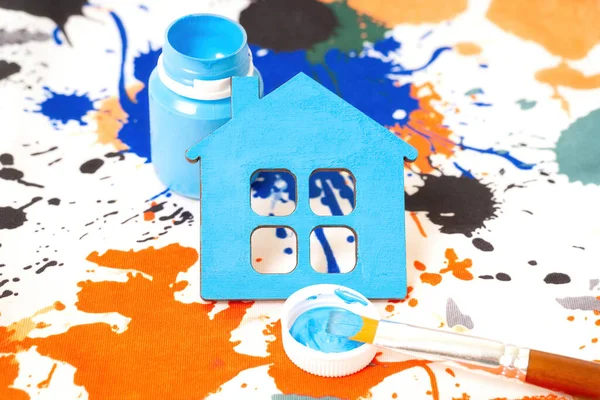 Freshly painted toy wooden house with paint bottle and brush on a watercolor fabric background.