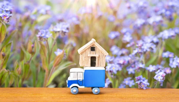 Toy blue vehicle carrying a small wooden house model against a blooming flowerbed. Creative home relocation solution concept.