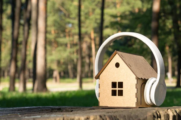 Toy plywood house placed on a tree stump in the forest having white wireless headphones on top. Finding peace in a rural area.