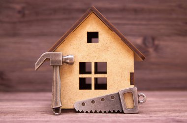 Minyatür kontrplak ev modeli, çekicin küçük bir kopyası ve tahta arka planda bir el testeresi var. Ev yapımı ve onarım konsepti.