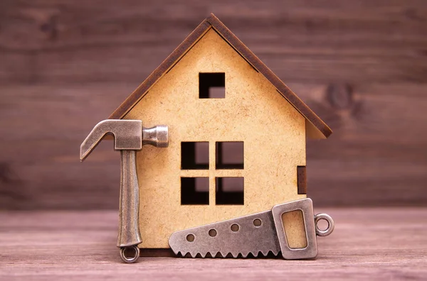Minyatür kontrplak ev modeli, çekicin küçük bir kopyası ve tahta arka planda bir el testeresi var. Ev yapımı ve onarım konsepti.