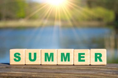 Word SUMMER made from wooden alphabet blocks against a lake background. Summertime vacation concept.