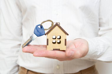 Minyatür ahşap ev modeli anahtarlığı olan ev anahtarını izleyiciye sunan bir adam. Gayrimenkul konsepti.