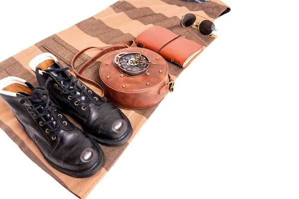 Clothes and accessories flat lay: circle clockwork leather bag, vintage work boots, journal, sunglasses, dress isolated on white.