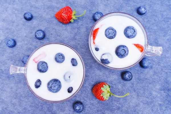 Two cups of berry yogurts with scattered berries on blue, top view. Summer seasonal dessert.