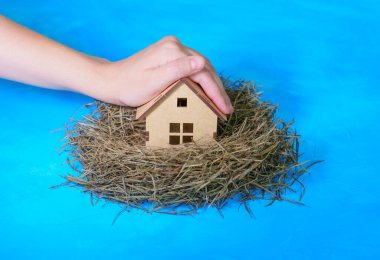 Hand covers a miniature wooden house placed in a hay nest isolated on blue background. Home insurance concept.