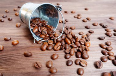 Bucket of dark roasted coffee beans scattered on a wooden background.