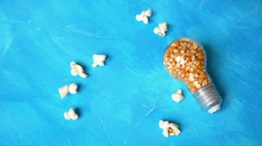 Incandescent light bulb full of popcorn kernels on a blue background. Popped corns falling down from above.