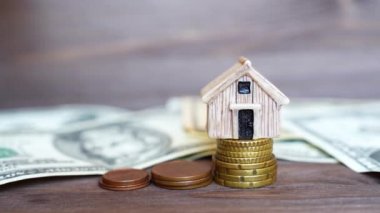 Hand puts a tiny key by a small toy house model placed on a coins stacks on a wooden table with dollar banknotes.