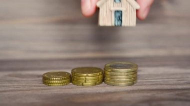 Hand puts a small house model on a coins stack and takes it back against a wooden background.