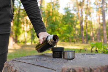 Turist termos şişesinden sıcak kahve dökerek ormandaki bir ağaç kütüğüne yerleştirilmiş iki fincana dolduruyor. Yürüyüş, seyahat ve aktif yaşam tarzı konsepti