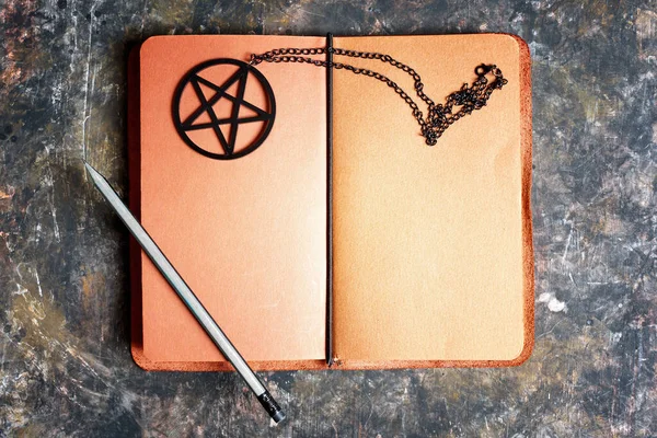 Brown leather notebook diary with pencil and black pentagram necklace on a dark grungy background. Creative spell book concept.