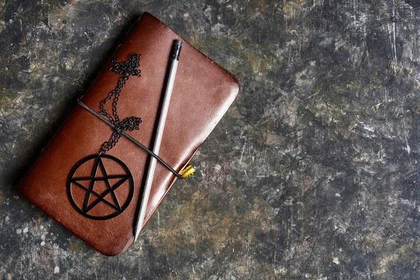 Brown leather pagan notebook with black pentagram and pencil on a dark grungy background. Personal spell book.