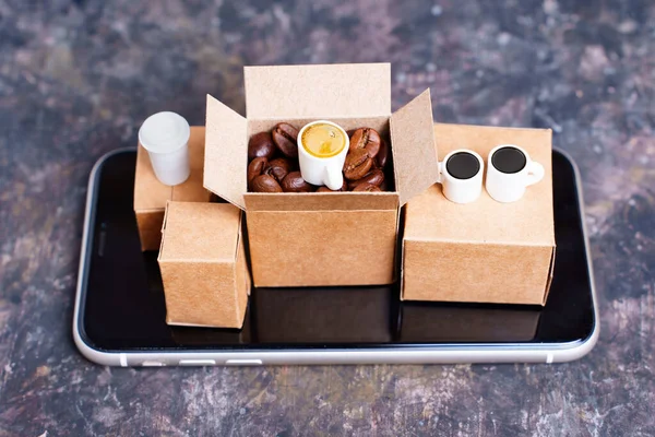 Miniature shipping boxes with roasted coffee beans and coffee cups placed on a smartphone isolated on a stained grungy background. Creative coffee roasting and supply concept.