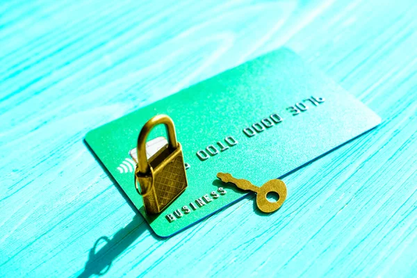 Close-up of a green credit card with bronze toned padlock and master key isolated on a blue wooden background, selective focus.