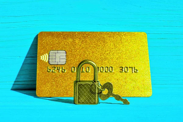 Keyed padlock in front of a golden credit card placed on a blue wooden background. Financial fraud protection concept.