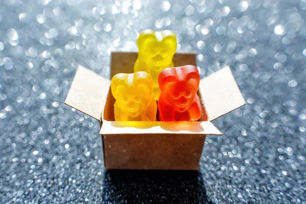 Close-up of three colorful bear shaped gummy candies in a tiny cardboard box against a dark glittering background.