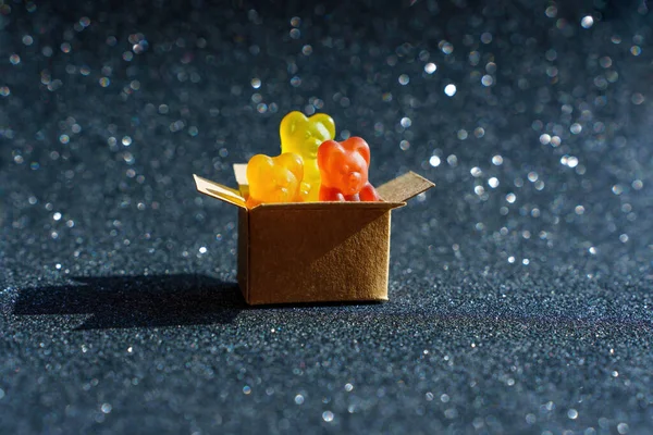 Three colorful gummy bears peeping out of a cardboard package isolated on black glittering background.