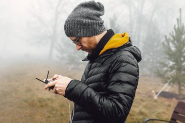 Sisli bir ormanda elinde İHA kontrol paneliyle duran bir adam. Bir kuadkopter uçuruyor..