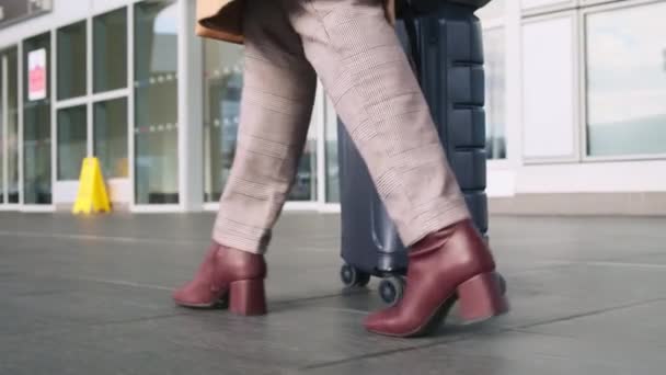 Vrouw in beige jas, lichte broek, kastanjebruine hielschoenen, met grijze koffer op wielen en tas, loopt buiten langs luchthaventerminal, kijkt naar haar telefoon. Reizen per vliegtuig concept, zakenreis — Stockvideo
