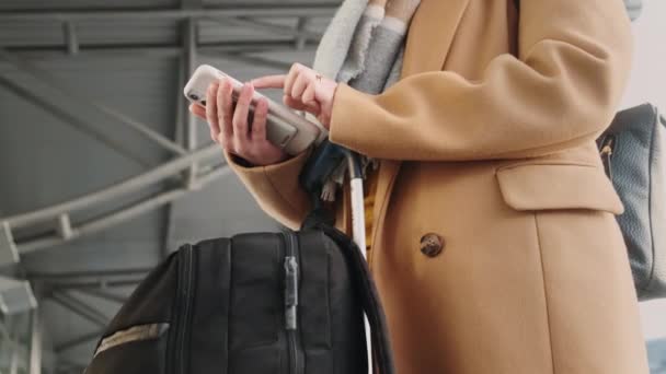 Vista lateral inferior de la mujer sin rostro con abrigo beige, bufanda de lana de pie dentro de la terminal del aeropuerto detrás de la bolsa de equipaje negro, surfeando en su teléfono con powerbank. Concepto de aplicación móvil. — Vídeo de stock