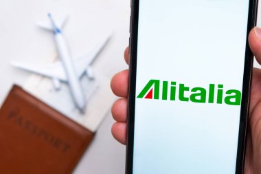 Alitalia Havayolları uygulaması akıllı telefon ekranının elinde. Beyaz oyuncak bir uçak ve pasaport, hafif bir yüzeyi olan bir masada yatıyor. Kasım 2021, San Francisco, ABD.